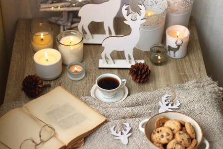 Various Christmas decorations, lit candles, cup of tea or coffee, cookies, books, presents and soft blanket on the table. Holiday hygge at home. Selective focus.の写真素材