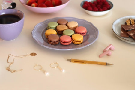 Plate of pastel macarons, cookies and chocolate, cup of tea of coffee, glass of bubble water, various berries, books and accessories on the table. Selective focus, pastel colors.の写真素材