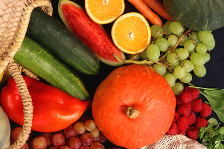 Round straw bag and various seasonal fruits and vegetables on dark background. Summer and fall produce. Top view.の写真素材