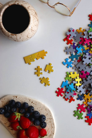 Colorful jigsaw puzzle pieces, cup of tea or coffee, eyeglasses and fresh berries for snacking. Hygge at home. Top view.の写真素材