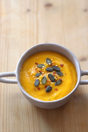 Bowl of creamy pumpkin and potato soup, with pumpkin seeds and chilli flakes toppings. Selective focus, wooden background.の写真素材