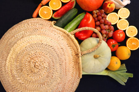 Round straw bag and various seasonal fruits and vegetables on dark background. Summer and fall produce. Top view.の写真素材
