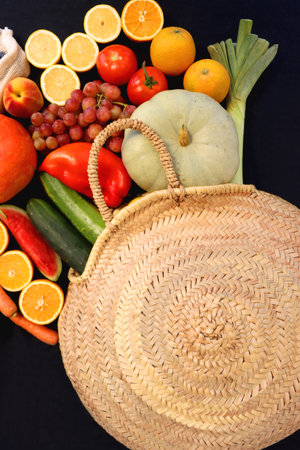 Round straw bag and various seasonal fruits and vegetables on dark background. Summer and fall produce. Top view.の写真素材