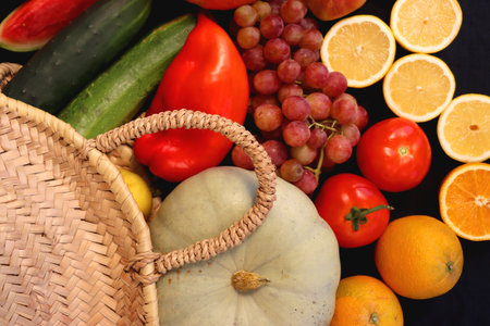 Round straw bag and various seasonal fruits and vegetables on dark background. Summer and fall produce. Top view.の写真素材