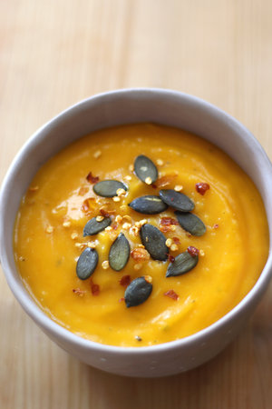 Bowl of creamy pumpkin and potato soup, with pumpkin seeds and chilli flakes toppings. Selective focus, wooden background.の写真素材