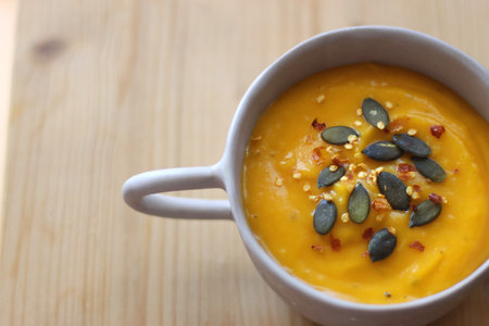Bowl of creamy pumpkin and potato soup, with pumpkin seeds and chilli flakes toppings. Selective focus, wooden background.の写真素材