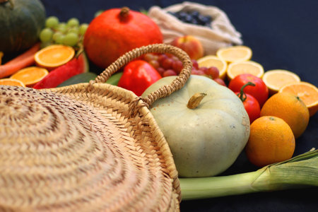 Round straw bag and various seasonal fruits and vegetables on dark background. Summer and fall produce. Selective focus.の写真素材