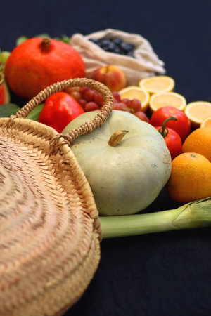 Round straw bag and various seasonal fruits and vegetables on dark background. Summer and fall produce. Selective focus.の写真素材