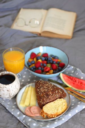 Tray with various healthy breakfast food, open book and reading glasses on the bed. Breakfast in bed concept. Selective focus.の写真素材