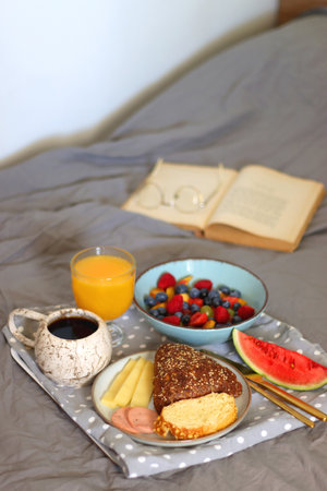Tray with various healthy breakfast food, open book and reading glasses on the bed. Breakfast in bed concept. Selective focus.の写真素材