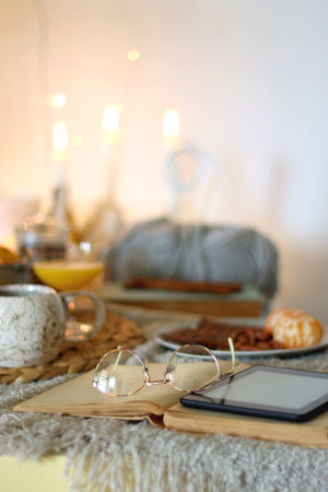 Books, e-reader, tea, orange juice, fruit, snacks, yarn, blanket and lit candles on the table. Autumn or winter hygge at home. Selective focus.の写真素材