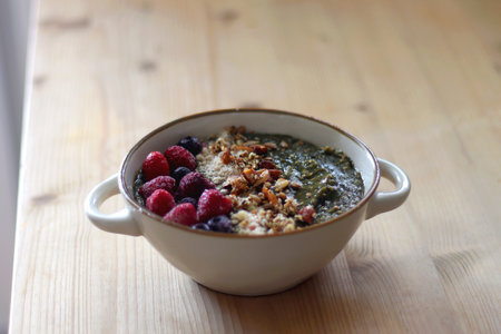 Beige bowl with green smoothie, frozen berries and homemade granola, served on the table. Selective focus.の写真素材