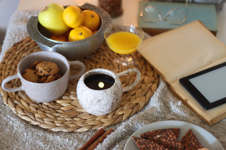 Books, e-reader, tea, orange juice, fruit, snacks, yarn, blanket and lit candles on the table. Autumn or winter hygge at home. Selective focus.の写真素材