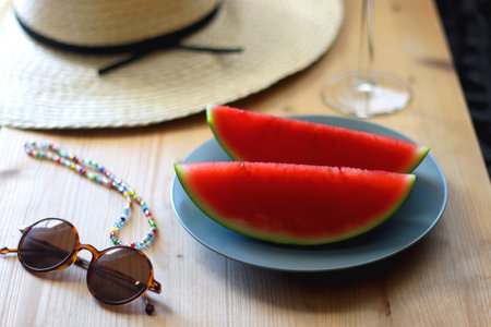 Plate with watermelon slices, glass of orange juice or cocktail, straw hat, colorful beaded necklace and sunglasses on the wooden table. Summer essentials concept. Selective focus.の写真素材