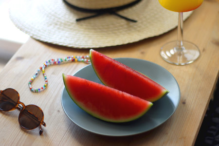 Plate with watermelon slices, glass of orange juice or cocktail, straw hat, colorful beaded necklace and sunglasses on the wooden table. Summer essentials concept. Selective focus.の写真素材