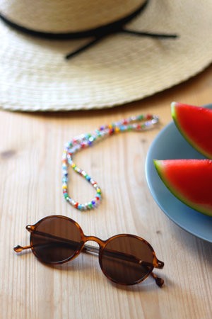 Plate with watermelon slices, glass of orange juice or cocktail, straw hat, colorful beaded necklace and sunglasses on the wooden table. Summer essentials concept. Selective focus.の写真素材