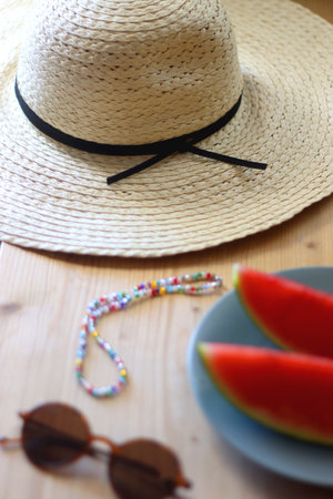Plate with watermelon slices, glass of orange juice or cocktail, straw hat, colorful beaded necklace and sunglasses on the wooden table. Summer essentials concept. Selective focus.の写真素材
