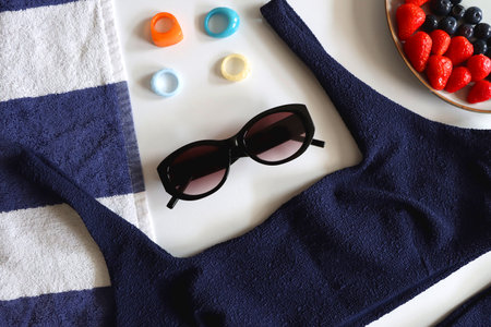 Navy swimsuit, striped beach towel, sunglasses, colorful rings and plate with berries. Top view.の写真素材
