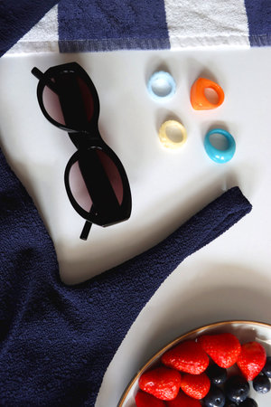 Navy swimsuit, striped beach towel, sunglasses, colorful rings and plate with berries. Top view.