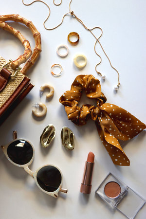 Wicker purse, sunglasses, scrunchie, lipstick and various gold and neutral jewelry on white...