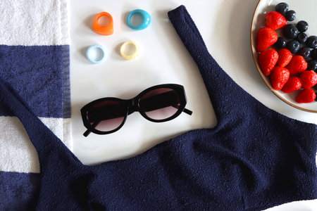 Navy swimsuit, striped beach towel, sunglasses, colorful rings and plate with berries. Top view.