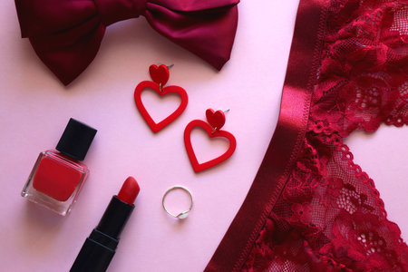Red lace bralette, hairbow, heart shaped earrings, golden ring, red lipstick and nail polish on pink background. Coquettish accessories for Valentine's day. Flat lay.の写真素材
