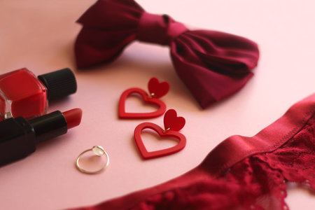 Red lace bralette, hairbow, heart shaped earrings, golden ring, red lipstick and nail polish on pink...