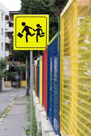 Yellow school zone street traffic sign, showing running boy and girl icons, next to a colorful school fence, on a bokeh daytime city streetの写真素材