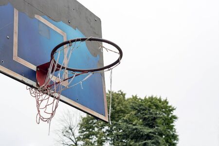 Torn old basketball hoop, on a grungy pealed off blue board, in a public park against the gray cloudy skyの写真素材