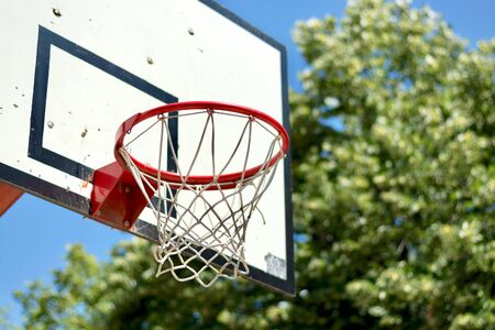Side view of red metal basketball hoop with white net and backboard, outdoors, against the green park trees and cler blue sky backgroundの写真素材