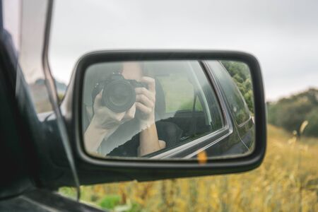 Blonde female professional photographer sitting in passenger seat taking a selfie in rear view mirror against bokeh background of misty yellow mountain meadow landscapeの写真素材