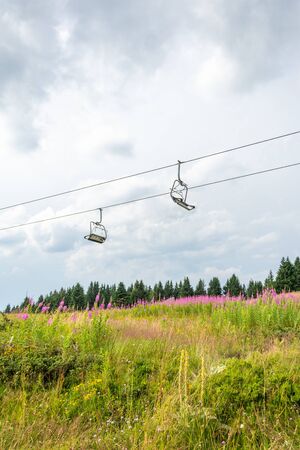 Two empty ski lift seats on cables above lush grassy summer mountain meadow covered with flowers and pine forest against cloudy skyの写真素材
