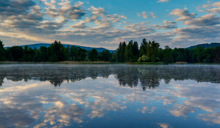 Sunrise on the pond in the mountains. Rudawy Janowickie. Clouds in the sky. Clouds are reflected in the water. Around the forest. Mountains in the background. Fog over the water.の写真素材