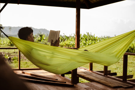 Asian man resting and reading a book in a comfortable hammock in a rural houseの写真素材