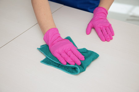 A hand in a pink glove wipes a white surface with a blue rag for cleaning