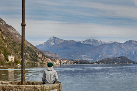 A young guy in a green hat is on the pier and is watching a beautiful view of the lake and the majestic mountains.のeditorial素材