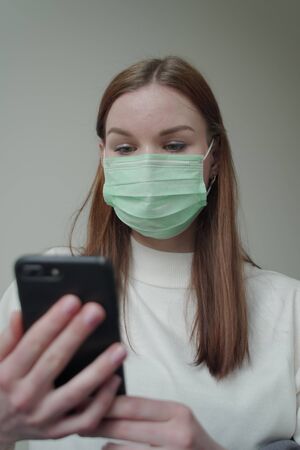 A girl in a medical mask holds a phone in her hands. Work on the phone during quarantine. Beautiful young girl with long red hair.の写真素材
