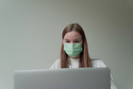 Young beautiful girl in a medical mask holds a laptop in her hands. Work from home during quarantine.の写真素材