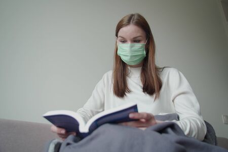 The young beautiful girl found an occupation during quarantine. Girl is reading a book in a medical mask. Orvi, coronovirus, influenza, acute respiratory infections.の写真素材
