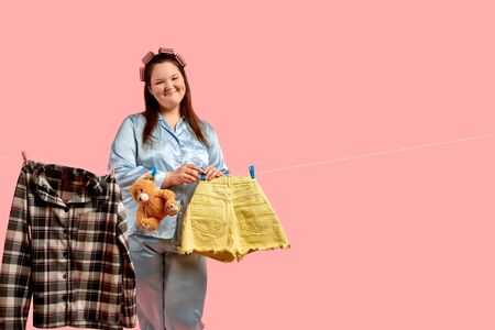 Cheerful beautiful fat girl in blue pajamas hangs clothes after washing, a good and happy wife. Pink background.の写真素材