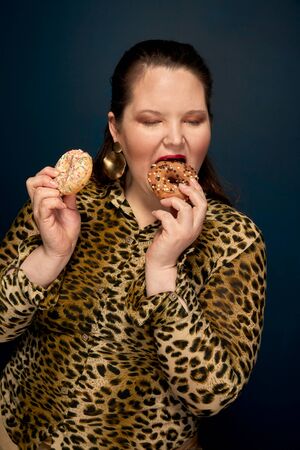 Fat girl eating donuts with an appetite. Leopard blouse. Overweight. Dark blue background.の写真素材