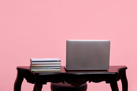 A white laptop is standing on a wooden table next to a stack of books on a pink background
