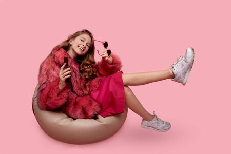 Beautiful young girl sitting in an armchair beige bag listens to music on the phone. Fashionable pink fur coat, pink dress and white sneakersの写真素材