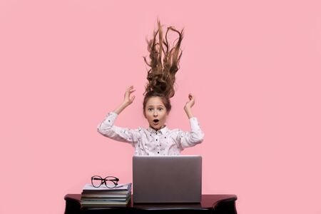 Beautiful young girl shakes with long blond hair and rejoices sitting at the computer. On a pink backgroundの写真素材