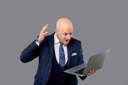 An angry bald man screams and raises his hand up. Looks at the screen. Laptop in hand. Grey background.の写真素材