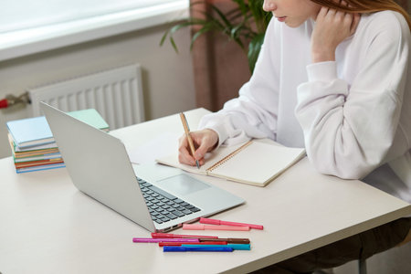 A young girl with long hair is studying online. Laptop on the table and felt-tip pens.