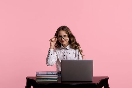 Young beautiful girl with loose long hair in glasses for vision sits at a wooden table with a laptop and a stack of booksの写真素材