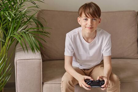 Young attractive guy sitting on a sofa holds a joystick. In the background is a green palm tree. He is full of happiness and victory.の写真素材