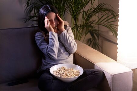 Girl is watching TV with a frightened face and popcorn on her lap. Sitting on the couch. A dark room. Cozy room.の写真素材