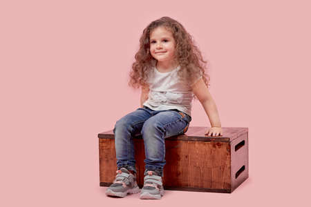 A small child in casual wear sits on a wooden stand on an empty background. The baby is blonde. Pink background.の写真素材